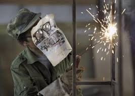 Homem a soldar com máscara e avental em ambiente industrial com faíscas