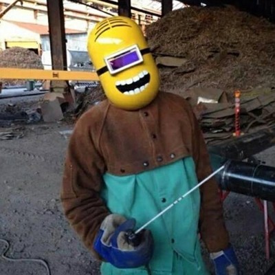 Pessoa com roupa de soldador verde e castanha, capacete amarelo com visor escuro e sorriso desenhado, em oficina industrial.