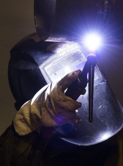 Pessoa a segurar tocha de soldadura acesa com máscara de soldador ao fundo