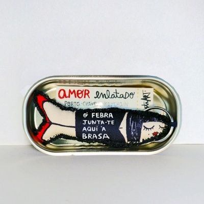 Lata metálica com porta-chaves em forma de peixe de tecido, cores preto, vermelho e bege, com texto em português.