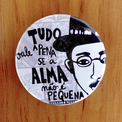 Botton redondo com texto e ilustração a preto e branco sobre fundo cinzento, em cima de madeira castanha