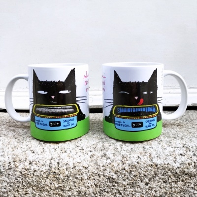 Duas canecas com ilustração de gato preto e latas de sardinhas, base verde, posadas em superfície de pedra.