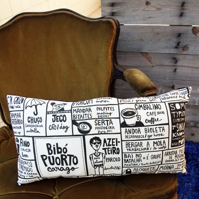 Almofada retangular preta e branca com desenhos e textos, sobre poltrona de veludo castanho