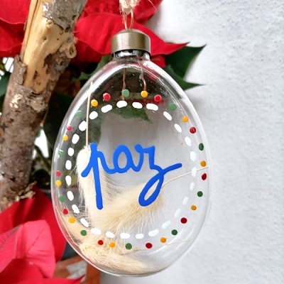 Ornamento oval transparente com a palavra paz e uma pena branca dentro, pendurado em ramo de árvore com folhas e flores vermelhas.