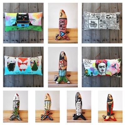 Almofadas e peixinhos decorativos em tecido com desenhos e textos variados, dispostos em fundo de madeira clara e parede cinza