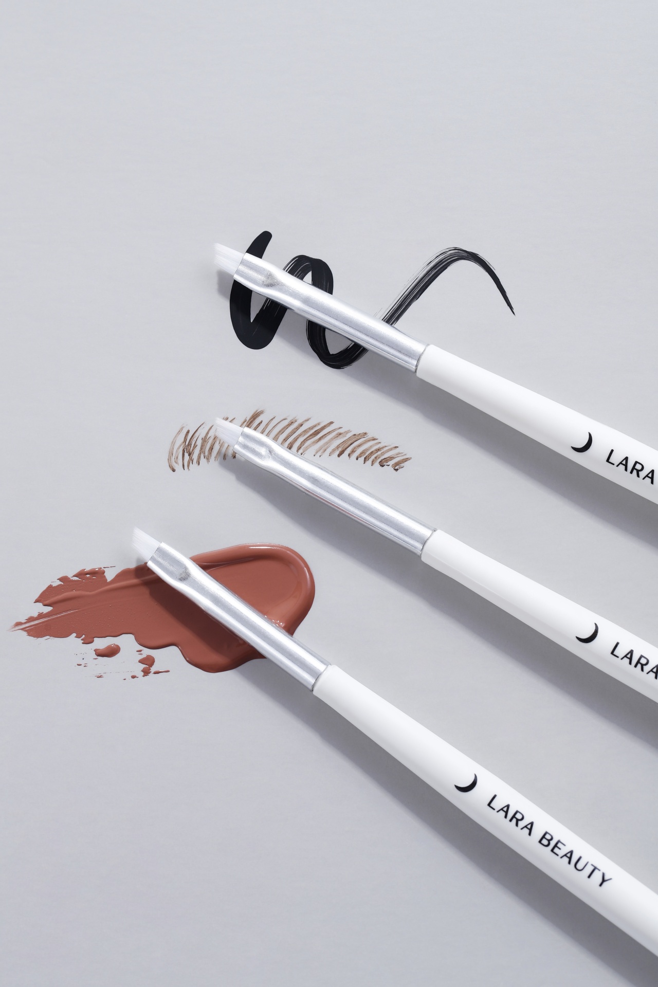 Pincel Chanfrado Lara Beauty Três pinceis de maquilhagem LARA BEAUTY com traços pretos, castanhos e líquido castanho-alaranjado sobre fundo cinzento.