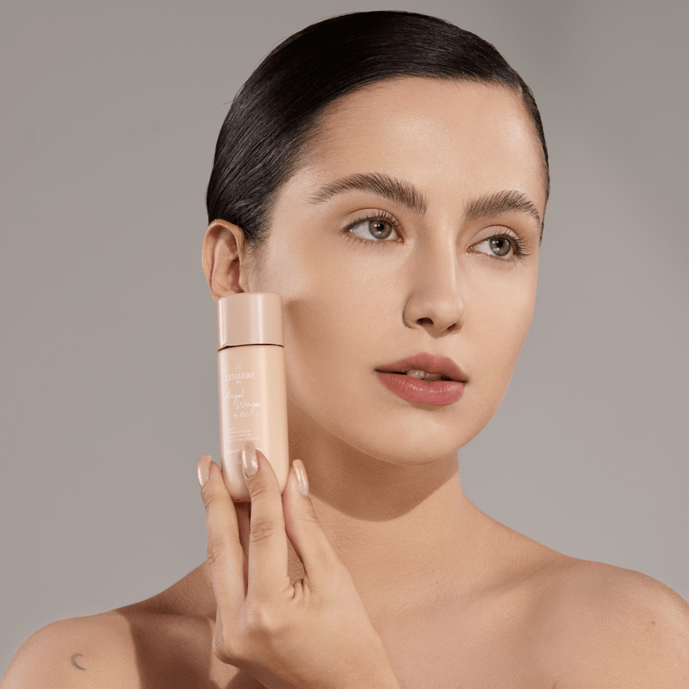 Base Alta Cobertura Angel Wings by Pri Lessa - Catharine Hill Mulher segurando frasco de creme facial bege claro com tampa rosada perto do rosto