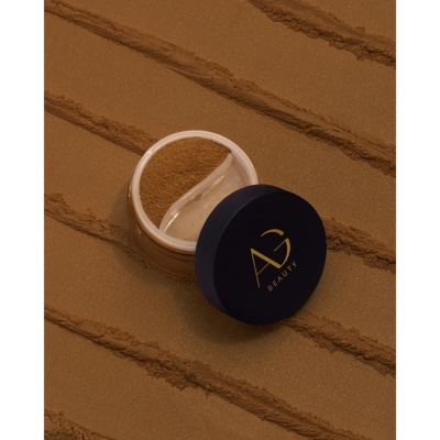 Pó facial solto em embalagem transparente com tampa azul e logo dourado sobre pó marrom