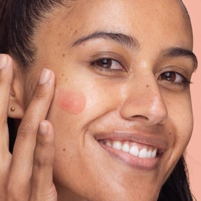 Rosto de mulher com creme rosa aplicado na bochecha em fundo pêssego