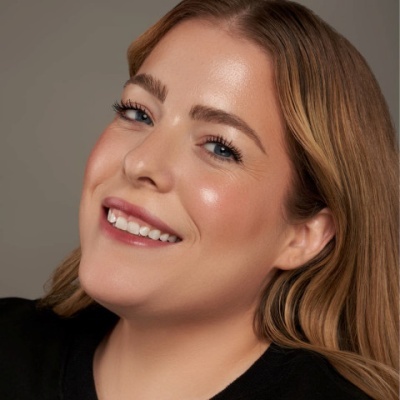 Mulher sorridente com cabelo louro e camiseta preta sobre fundo cinza.