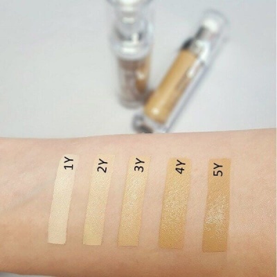 Braço com cinco tons de base líquida numerados 1Y a 5Y aplicados, com dois frascos da base ao fundo.