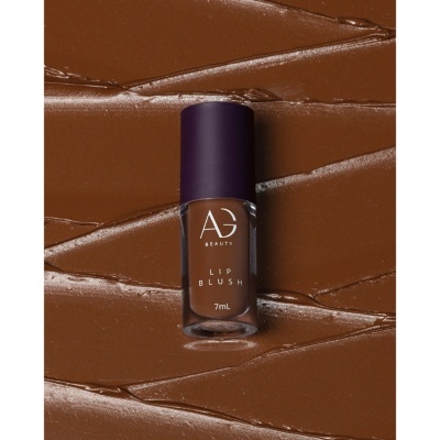 Blush labial castanho da AG Beauty em frasco de vidro com tampa roxa sobre fundo castanho líquido texturizado.