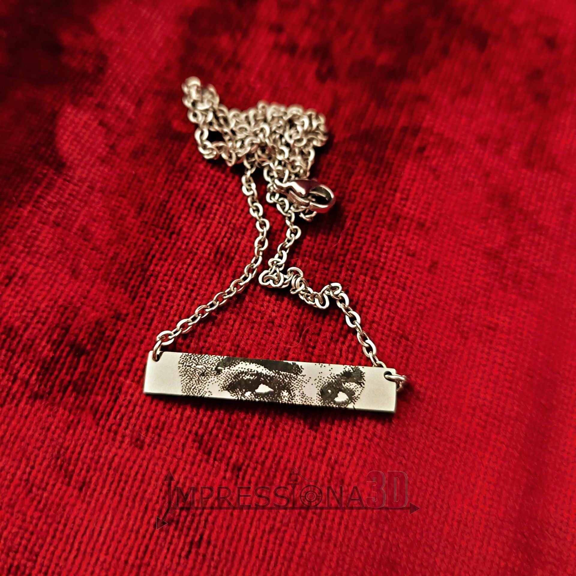 Colar prateado com pendente retangular estampado com olhos, sobre fundo vermelho