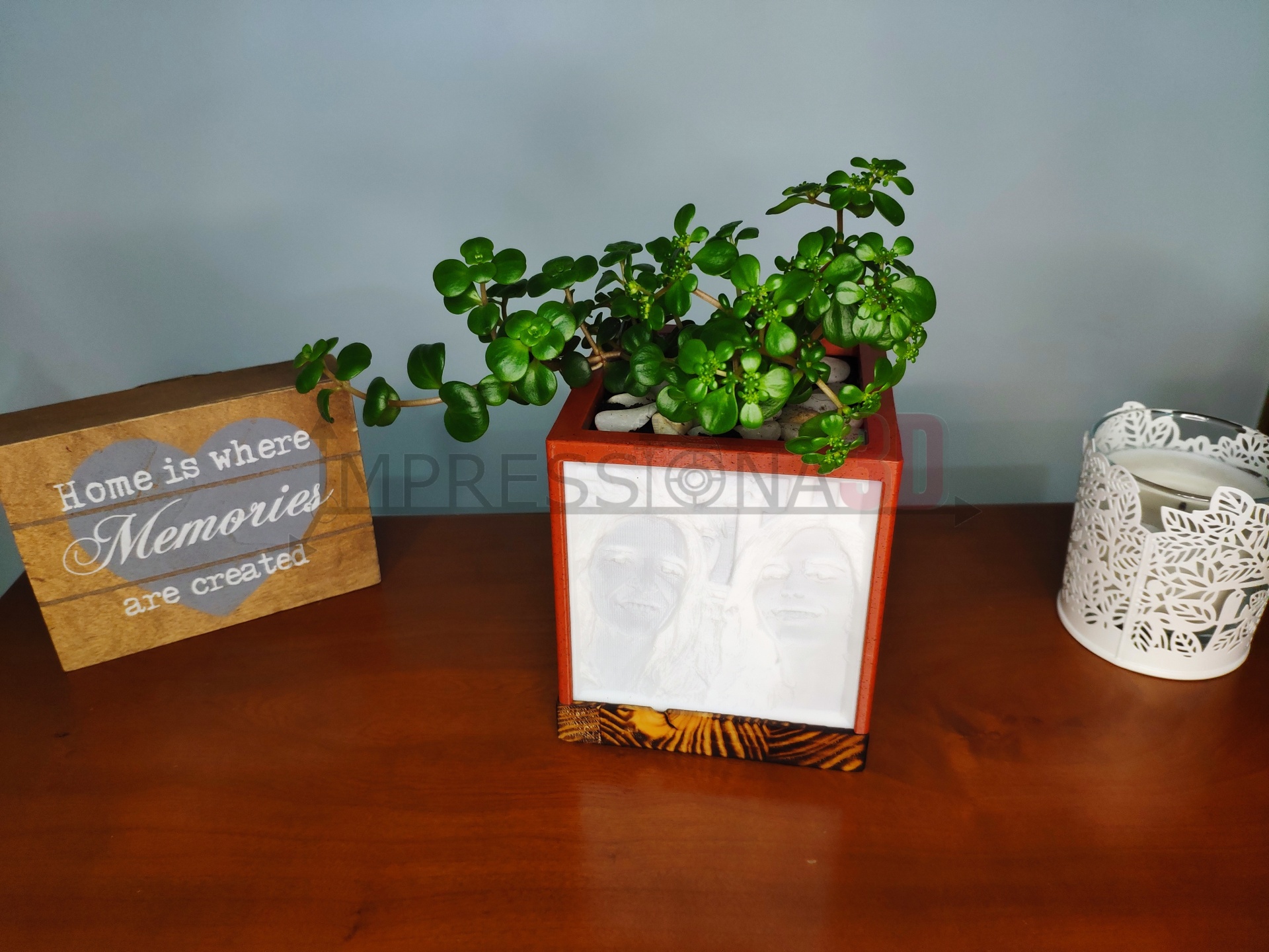Vaso de planta quadrado vermelho com planta verde sobre mesa de madeira, placa decorativa com texto e castiçal branco