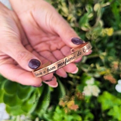 Pulseira metálica cor cobre com gravação em português segurada por mão com unhas roxas, fundo de plantas verdes.