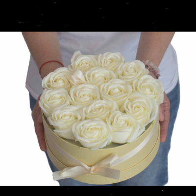 Caixa redonda creme com rosas brancas e laço, segurada por pessoa com camiseta branca e calças jeans