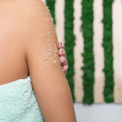 Braço com esfoliante e toalha verde-água, fundo com plantas verdes