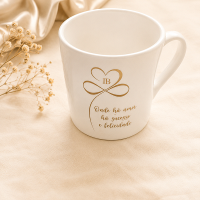 Caneca branca com texto e desenho em dourado, sobre superfície creme com tecido e flores secas