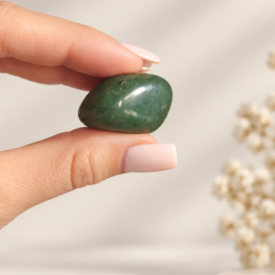 Mão segurando uma pedra verde polida, fundo neutro com decoração de flores secas