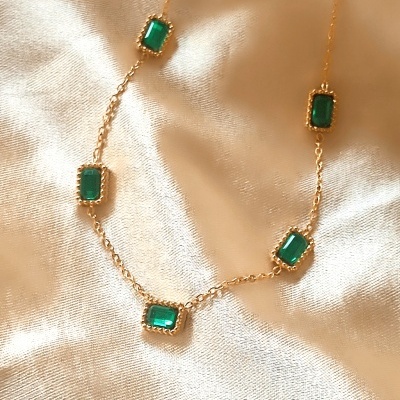 Colar dourado com pedras verdes retangulares sobre tecido creme.