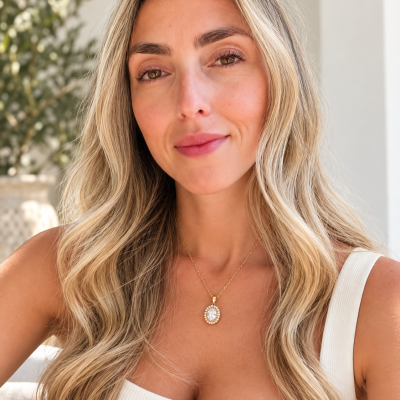 Colar com pendente oval dourado com pedras transparentes no pescoço de mulher loira com vestido branco