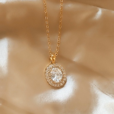 Colar dourado com pendente ovular e pedras transparentes sobre tecido bege