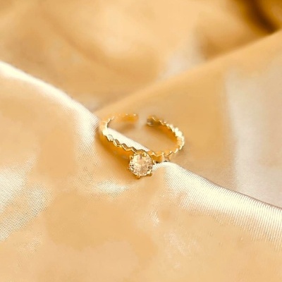 Anel dourado com pedra transparente sobre tecido acetinado bege.