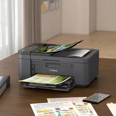 Impressora Canon a imprimir documentos coloridos numa mesa de madeira com papéis e smartphone.