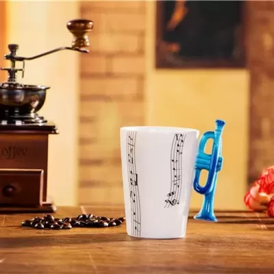 Caneca branca com notas musicais e pega azul em forma de trompete ao lado de moinho de café e grãos sobre mesa