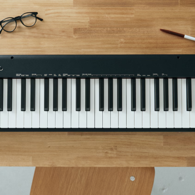 Teclado musical digital preto sobre mesa de madeira com óculos, caneta, caneca e plantas