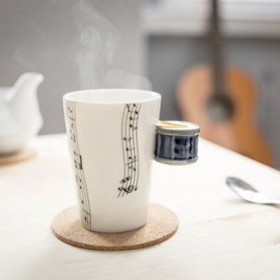 Caneca musical branca com notas pretas e pega preta com dourado sobre descanso de cortiça em mesa clara