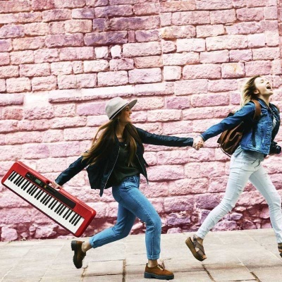 Duas mulheres jovens correndo com um teclado musical contra uma parede de tijolos rosa.