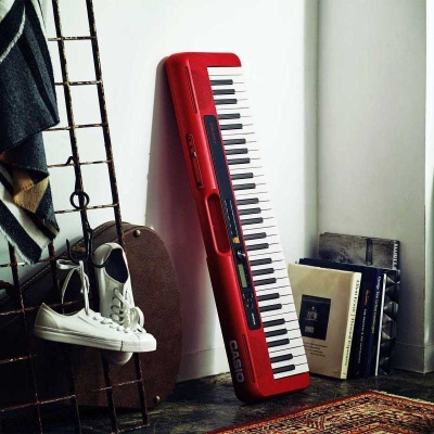 Teclado musical Casio vermelho com 61 teclas apoiado na parede junto a livros e sapatos.