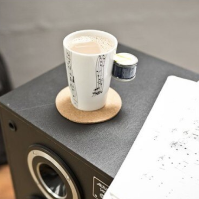 Caneca branca com notas musicais, bebida quente, base de cortiça, perto de partitura musical