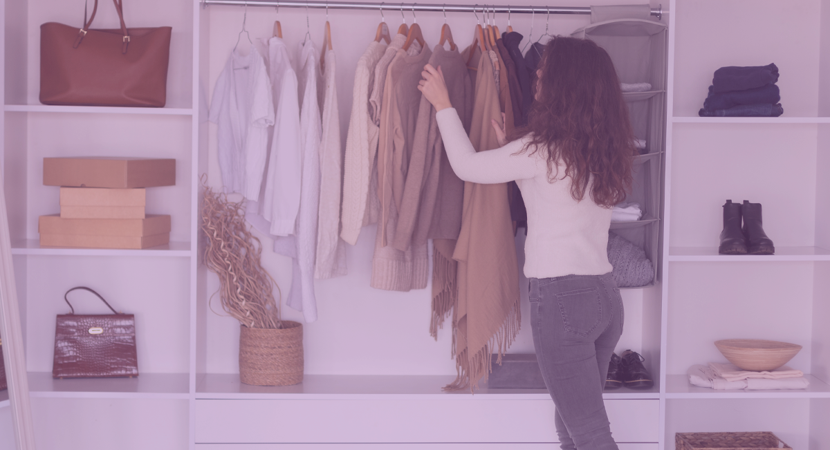 Armário aberto com roupas castanhas e brancas penduradas e prateleiras com caixas, malas e sapatos, mulher a escolher roupa