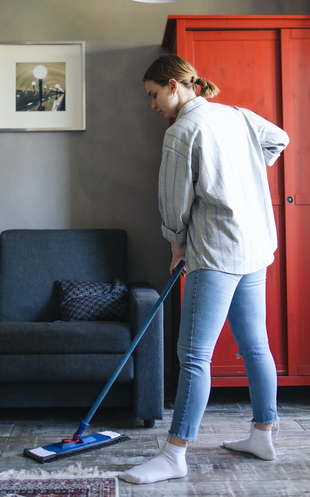 Mulher a limpar chão de madeira com mop numa sala com sofá cinza e armário vermelho