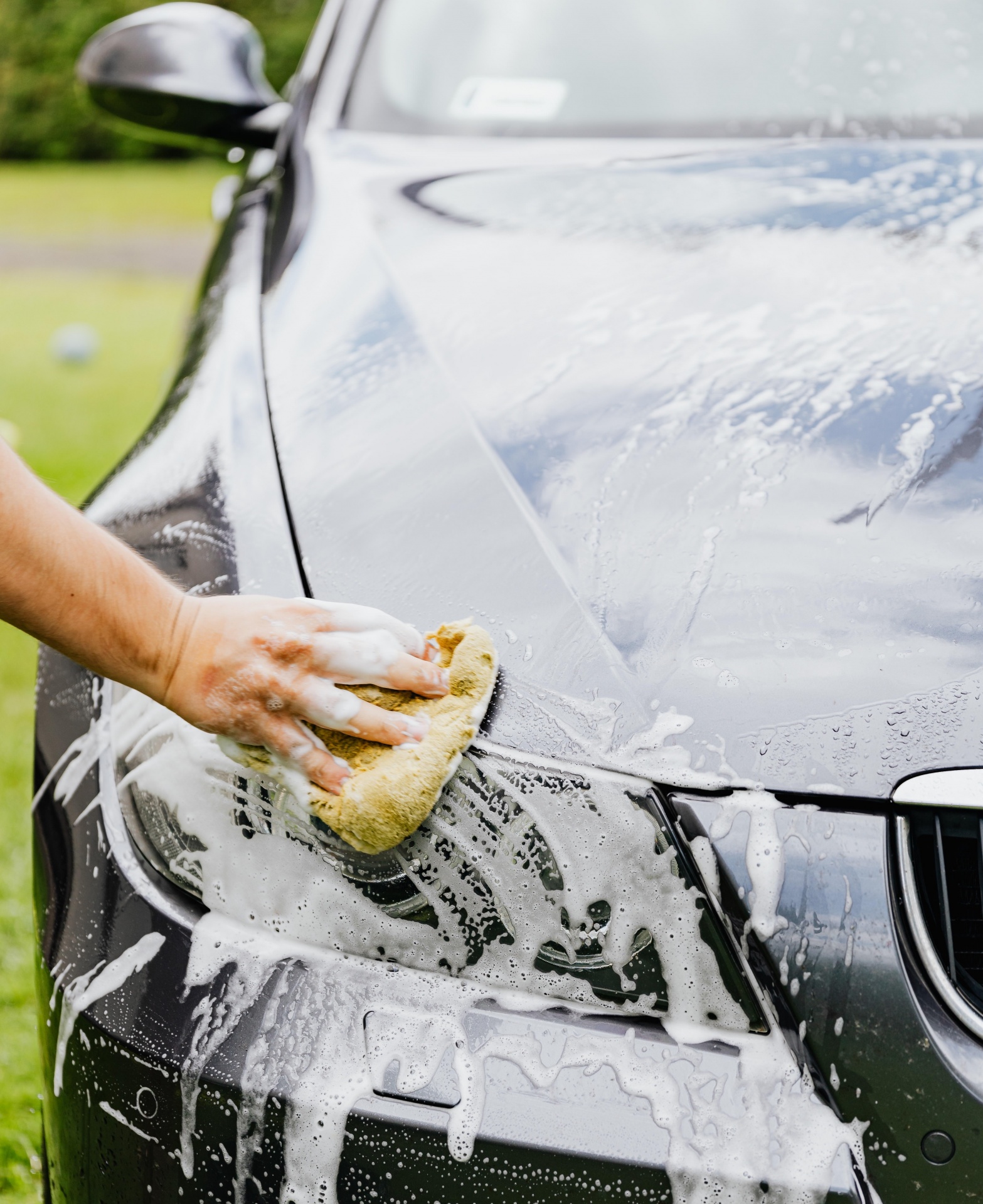 Carro preto com espuma branca a ser lavado com uma esponja amarela na mão