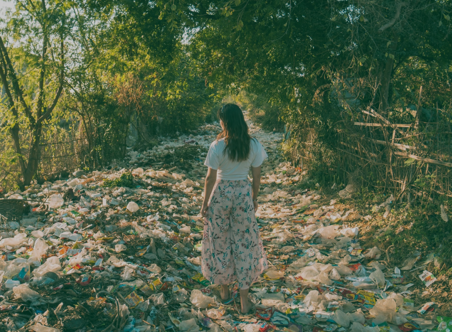 Mulher de costas com saia floral e camisola branca num caminho cheio de lixo plástico entre árvores