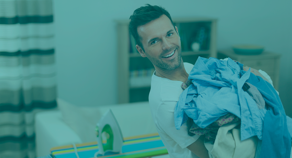 Homem segurando roupas coloridas em ambiente doméstico com ferro de engomar ao fundo