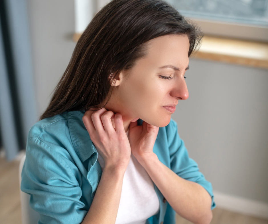 Mulher com expressão desconfortável a tocar a nuca, vestindo camisa azul e t-shirt branca, junto a uma janela