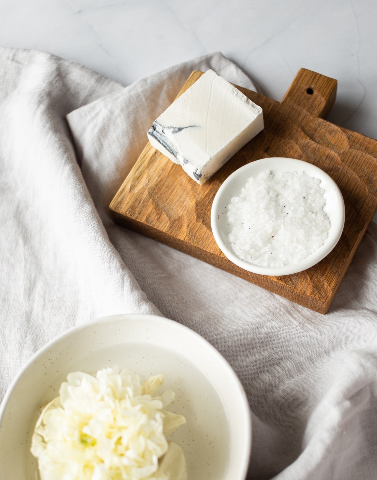 Tábua de madeira com cubo branco e prato de sal sobre pano bege, com taça branca com flor branca ao lado.
