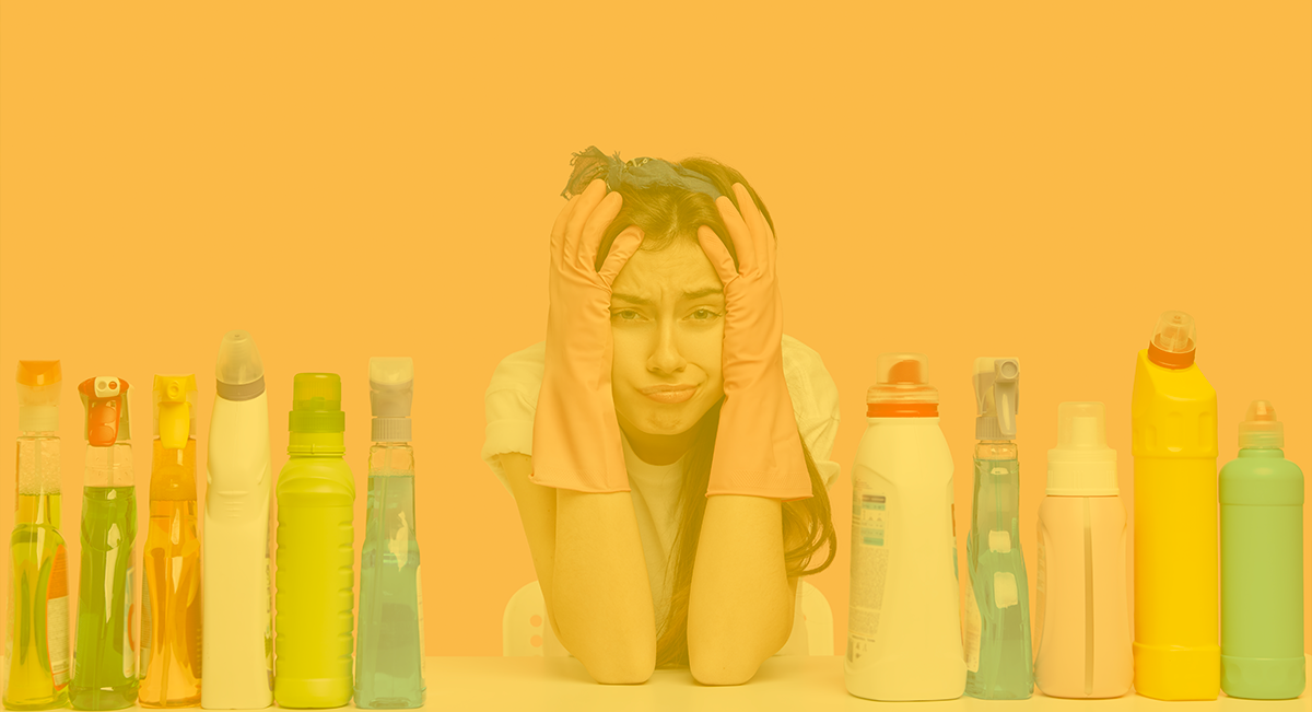 Mulher com luvas de borracha entre várias garrafas de limpeza coloridas sobre mesa com fundo amarelo