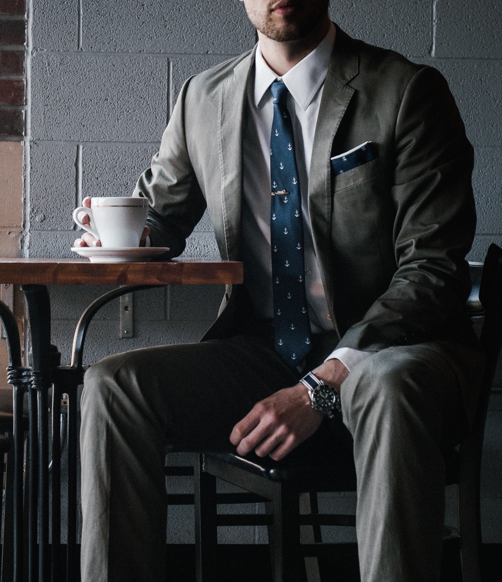 Homem sentado com fato cinza esverdeado, gravata azul e camisa branca numa mesa de café