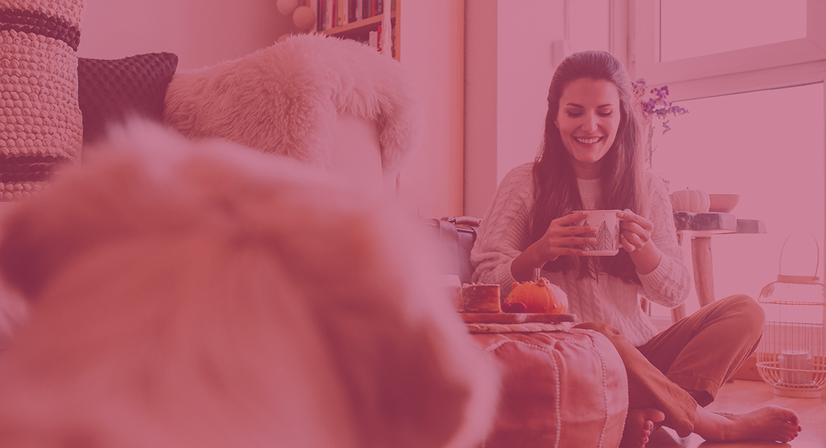 Mulher sorridente sentada no chão a segurar uma chávena num ambiente acolhedor de sala com almofadas e mesa com comida