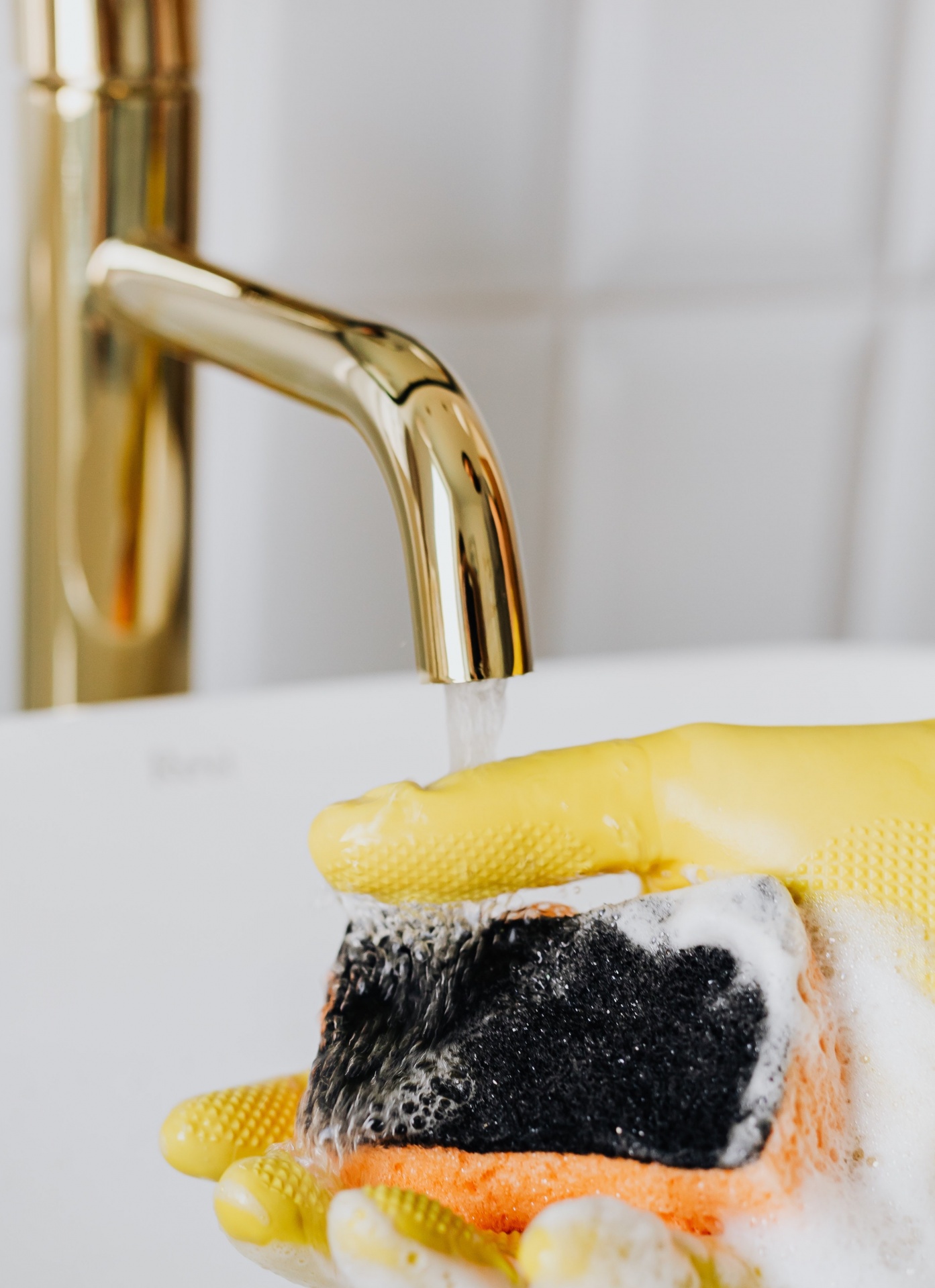 Mão com luva amarela segurando esponja com espuma debaixo de torneira dourada