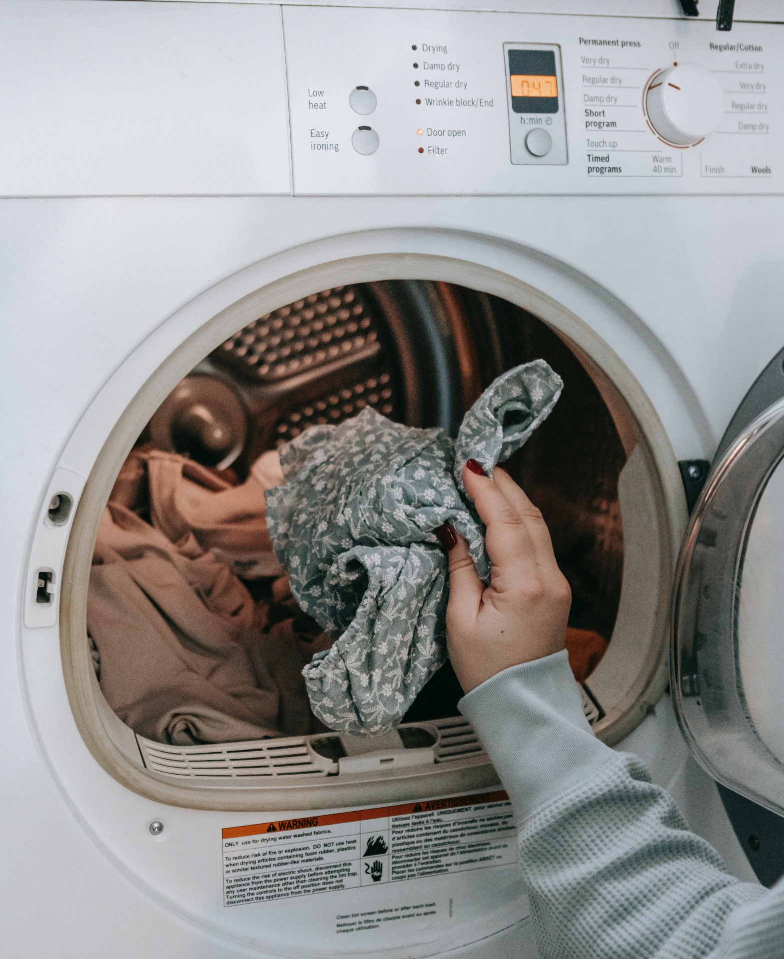 Máquina de secar roupa branca aberta com roupa dentro e mão segurando tecido floral