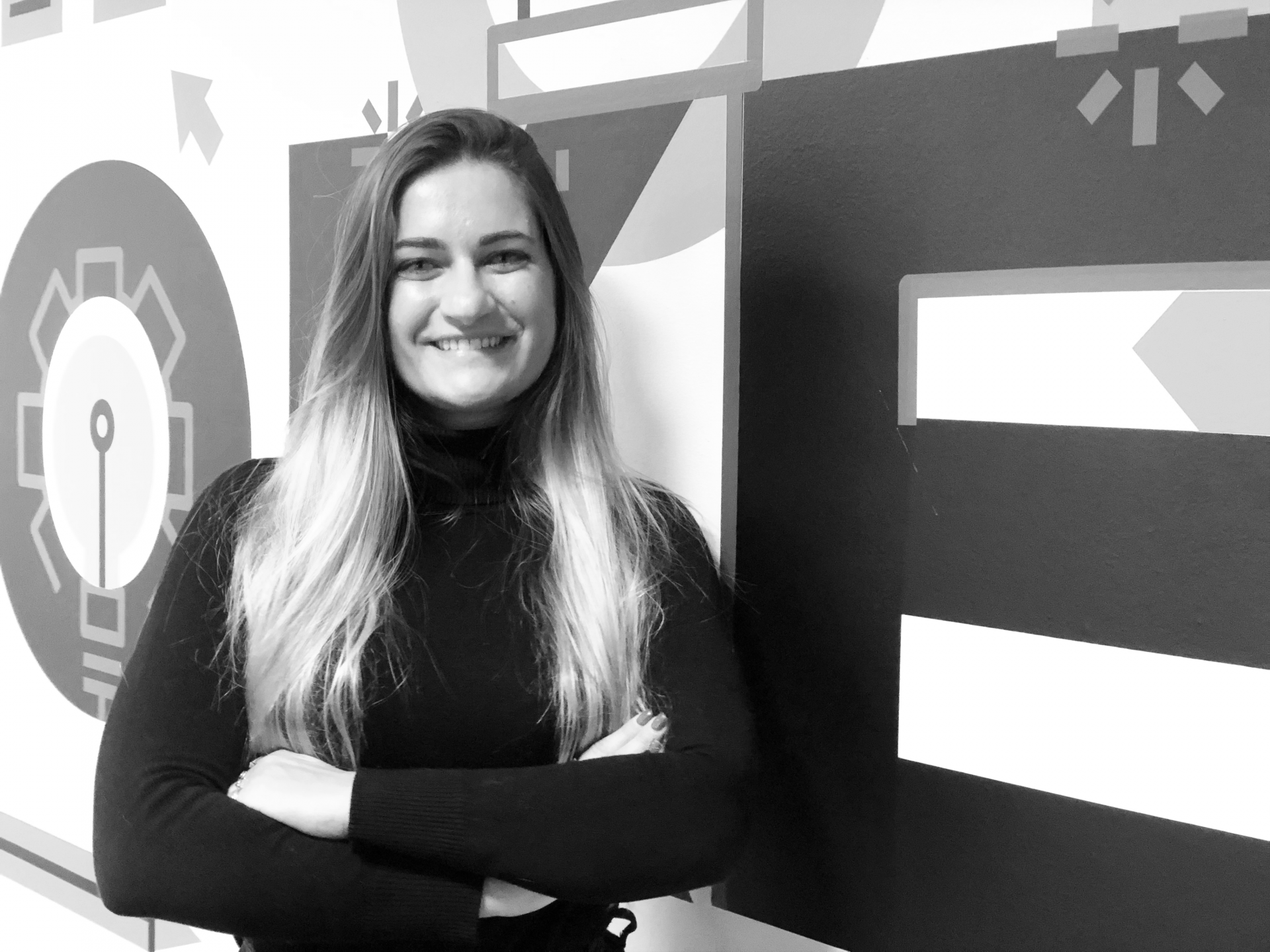 Mulher com camisola preta e cabelo comprido, em frente a parede com gráficos geométricos