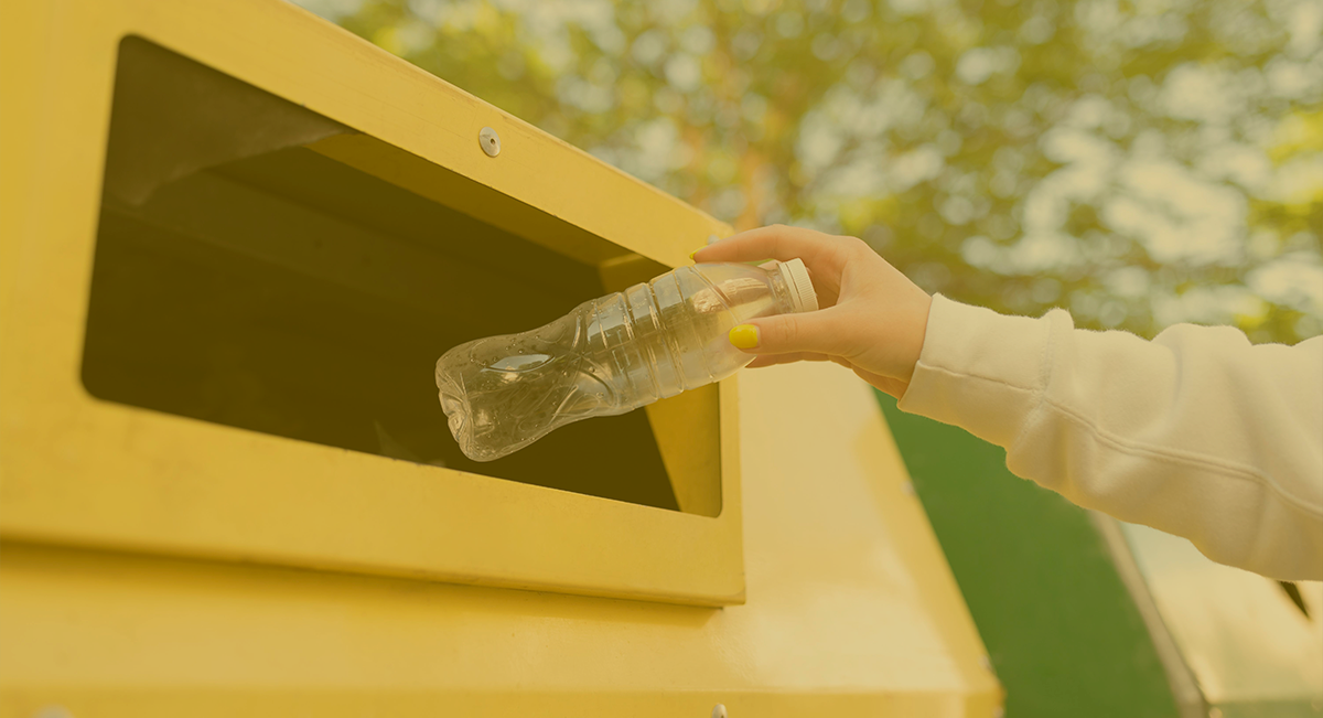 Pessoa a colocar garrafa plástica num contentor amarelo para reciclagem em ambiente exterior.