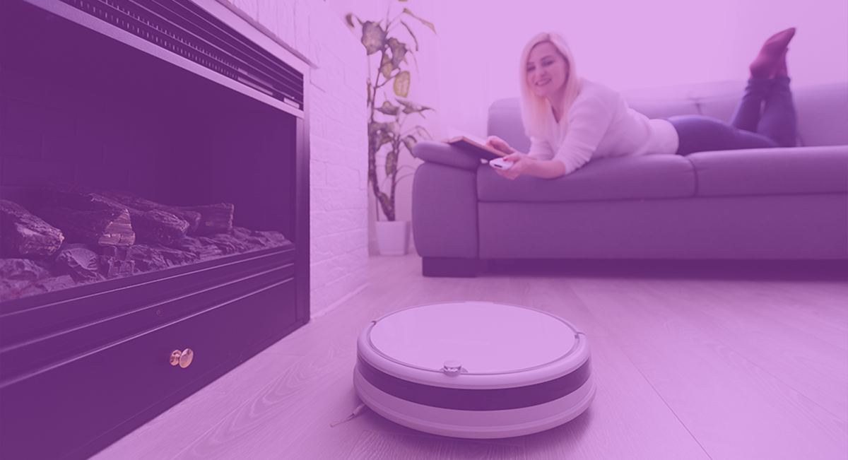 Robot aspirador circular branco e cinza em chão de madeira com mulher no sofá e lareira