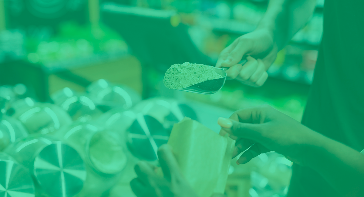 Pessoa a colher pó verde de um recipiente para um saco de papel castanho num mercado